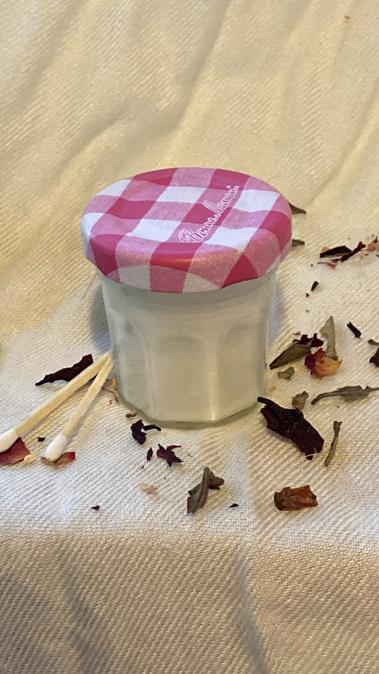 A mini jam jar candle with a pink gingham lid screwed on top. It's set against a silky white background and posed with two white matches and some dried tea leaves scattered around it.