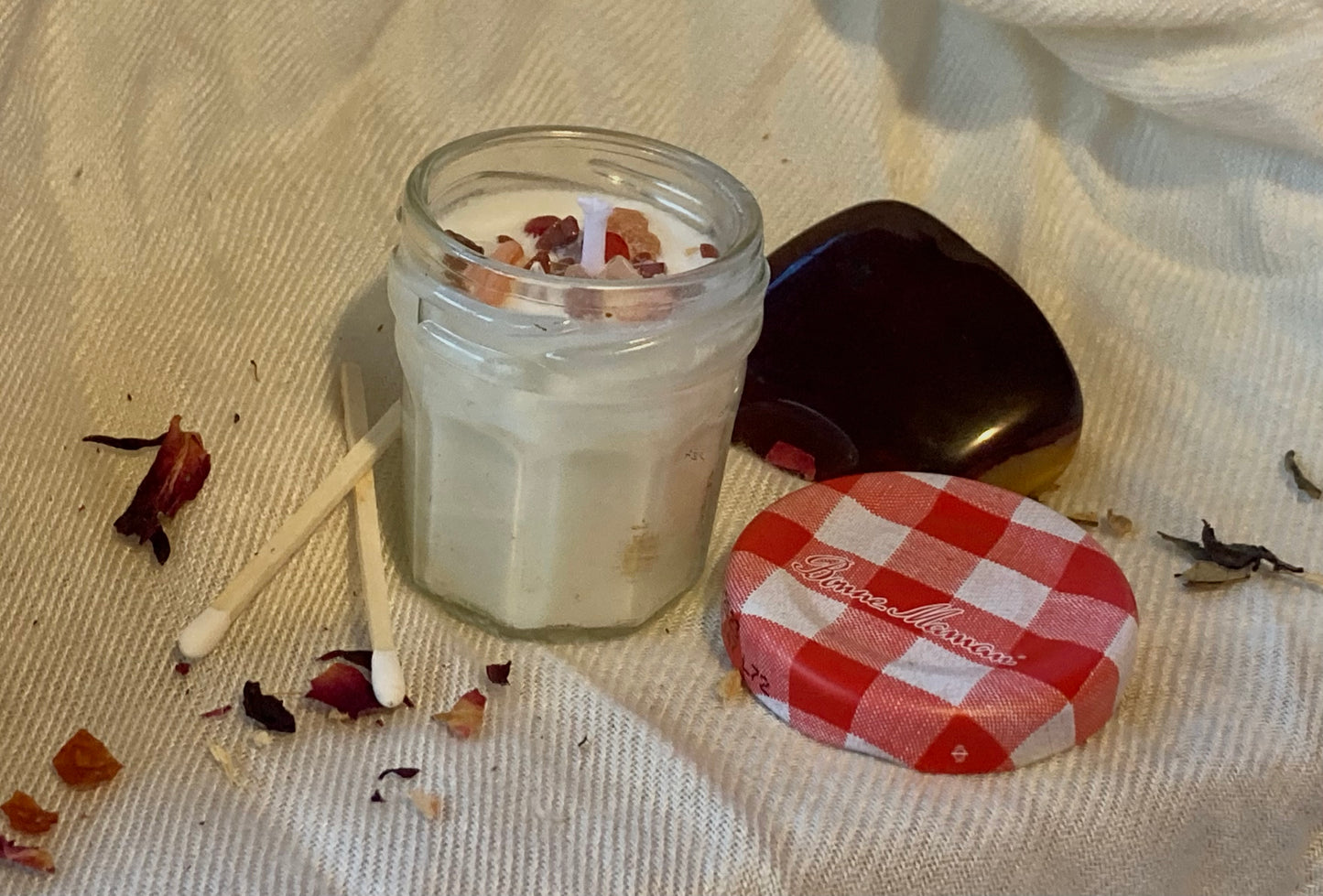 A jam jar mini candled topped with orange, red, and glittery brown crystal chips is centered against a silky white background. Two white matches are found on the left, while a large red and yellow carnelian stone and the red gingham-patterned lid of the mini jam jar are posed to the right of the candle.