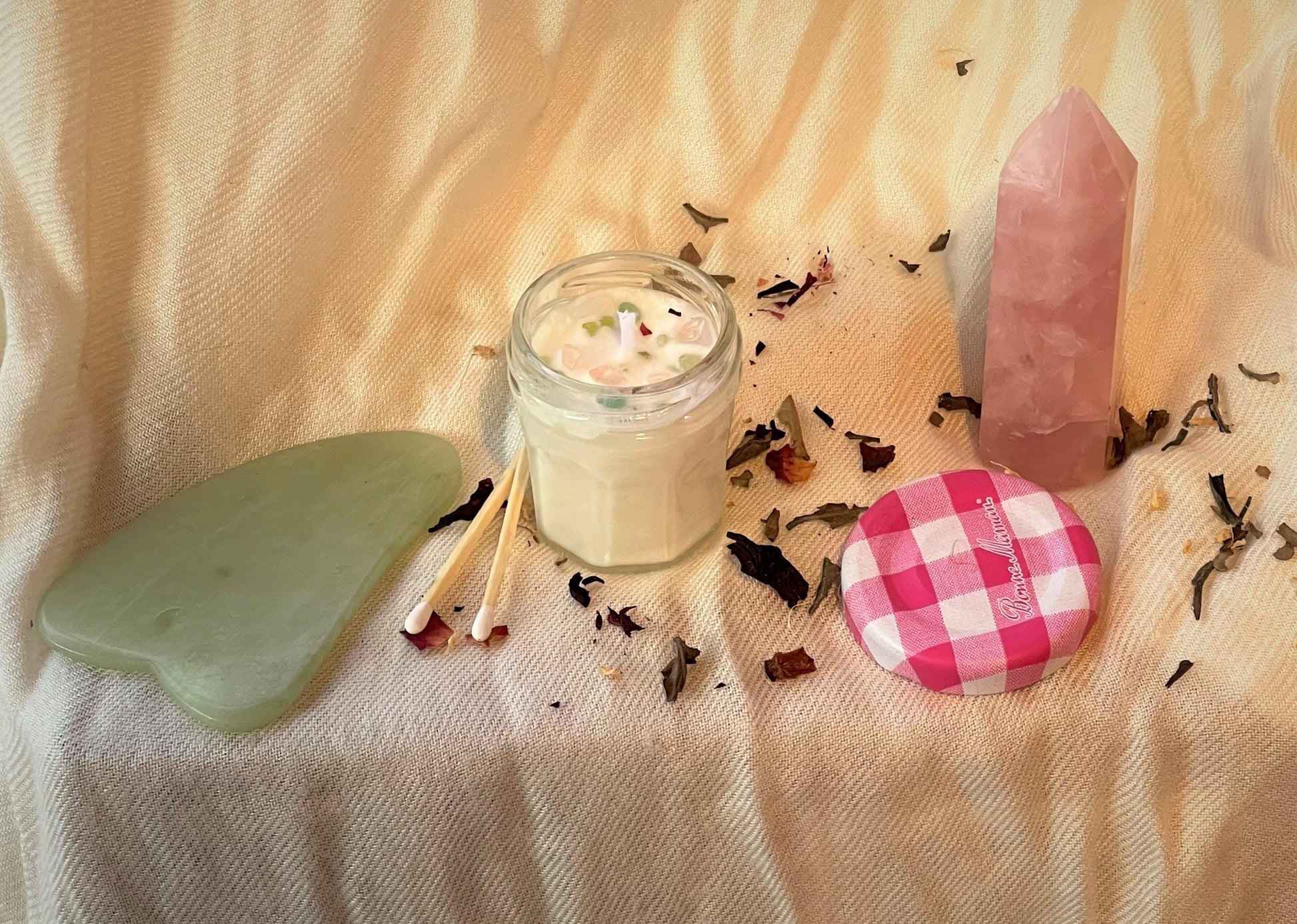 A mini jam jar candle topped with pink and green crystal chips sits centered in a silky white background. To the left, there is a green jade gua sha tool and two white matches. To the right, there is a rose quartz tower and the pink gingham candle lid. Dried tea leaves and rose petals are scattered among everything.